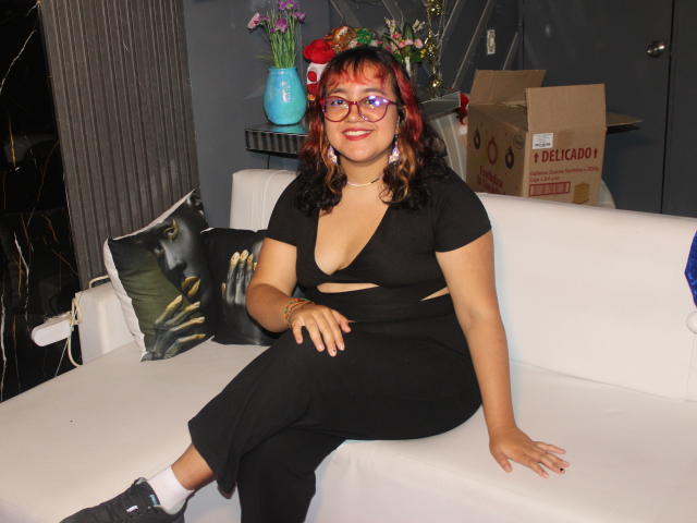NamiNerona, with red and black hair, glasses, and a low-cut black dress, sits on a white couch, showing her tits, wearing black shoes and white socks.