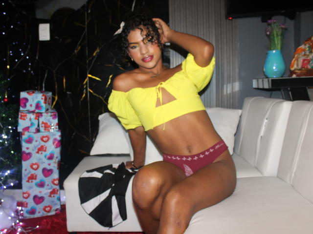 SamaraDuvall, a dark-skinned woman with curly black hair, sits on a white couch. She wears a yellow off-shoulder crop top with a triangular cutout and maroon panties with star patterns. Her right arm is raised, touching her head. The background includes a black wall with gold patterns, a blue vase with flowers on a shelf, and wrapped gifts with colorful patterns. Her relaxed, slightly tilted posture and confident expression are key elements. The image has a casual, festive atmosphere.