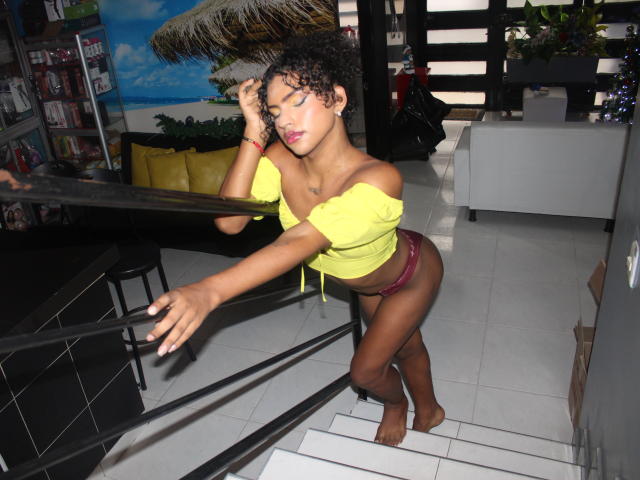 SamaraDuvall with curly hair, yellow off-shoulder top, and red lace panties, leaning on a black bar, eyes closed, hand in hair, standing on white tile stairs.