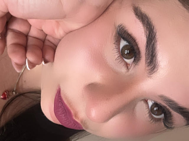 VenusxMusa's face with brown eyes, thick eyebrows, and purple lipstick. She's touching her cheek with her right hand, showing off a red bead necklace. Her skin is light.
