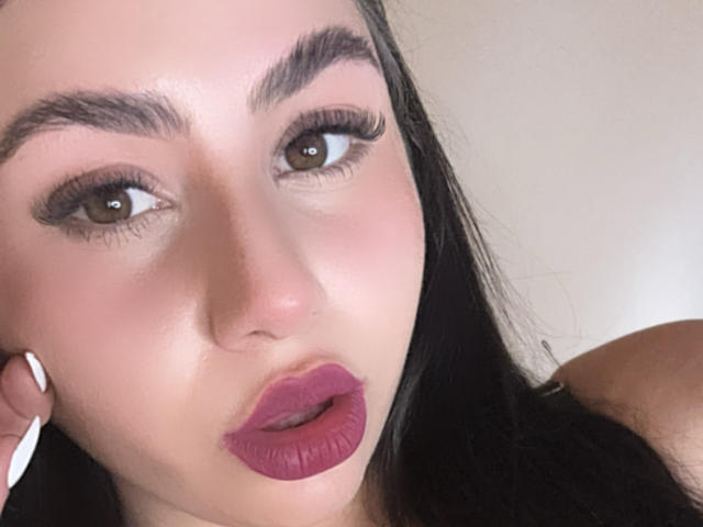 VenusxMusa has dark eyebrows, brown eyes, long black hair, white nails, and purple lipstick. Her expression is slightly open-mouthed. She has a light skin tone.