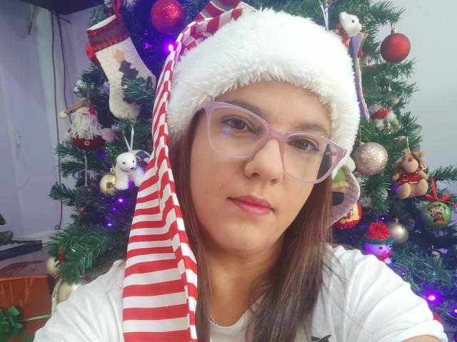 AmeliaWilson with pink glasses, red and white striped Santa hat, white fluffy trim, pink lipstick, brown hair, white shirt, standing in front of a decorated Christmas tree.