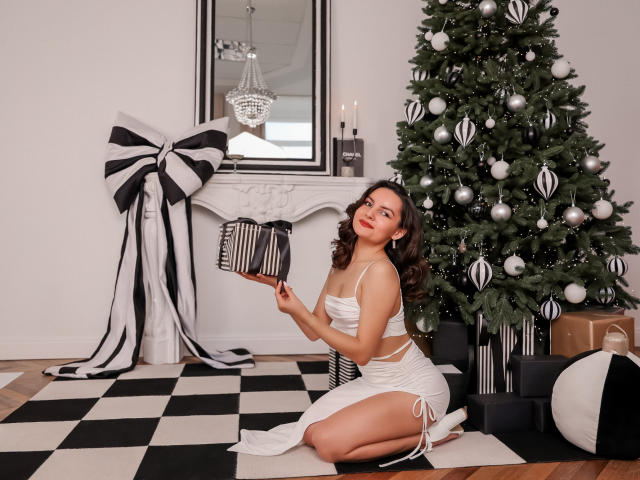 JasmineAsha, kneeling, wearing white strappy lingerie, holding a striped ornament, next to a decorated Christmas tree, with black and white gifts, and a large black and white bow.