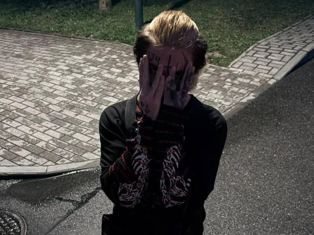BluesBruce with bleached blonde hair, tattoos on hands, covering face with hands, wearing black shirt with red and white pattern, standing on wet, dark asphalt, looking sad.