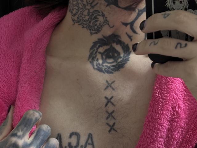 BluesBruce's tattooed chest with a pink robe, black rose tattoo, cross tattoos, and a "13" tattoo, holding a phone. Visible tattoos on neck and hand.