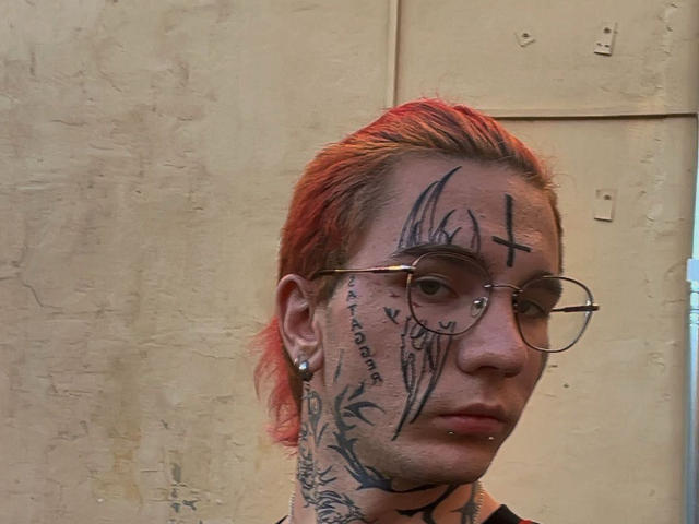 BluesBruce has red-dyed hair, tattoos on the face and neck, and wears glasses with a nose piercing and ear studs. Textured beige wall in background.