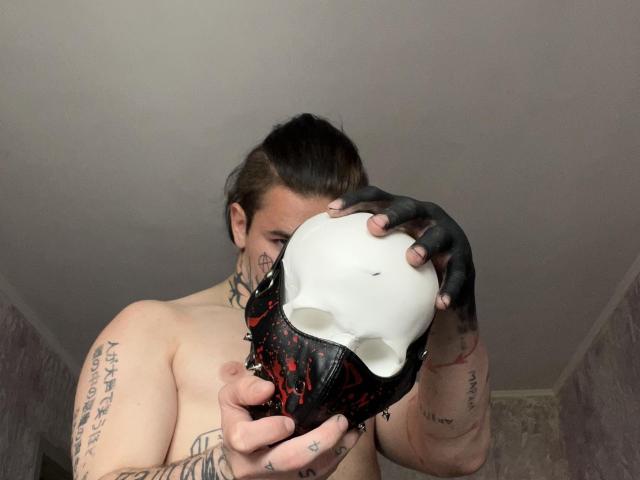 BluesBruce with tattoos holding a black and white skull mask, wearing black fingerless gloves, looking at the camera, shirtless, with brown hair in a ponytail.