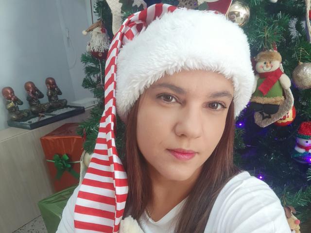 Amelia Wilson, con cabello castaño, usando un gorro de Papá Noel a rayas rojas y blancas, una camisa blanca y lápiz labial rosa, está de pie frente a un árbol de Navidad con adornos..