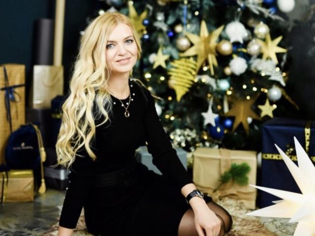 MelissaTyler with long blonde hair, black dress, sitting by a Christmas tree, smiling, wearing a black necklace, and a black watch, surrounded by gold and blue ornaments.