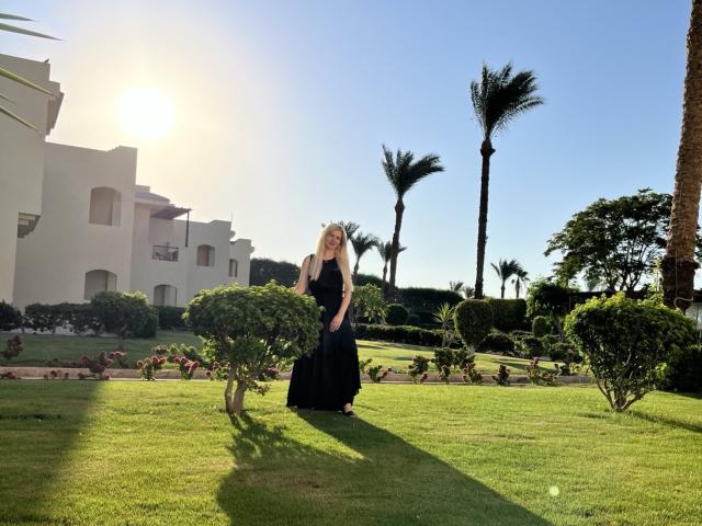 MelissaTyler with long blonde hair, wearing a black dress, standing on green grass, sun behind her, palm trees, white building, and small bushes in the sunny background.