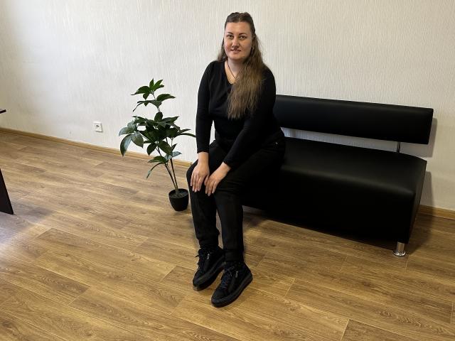 MonaLia with long brown hair, black long-sleeve shirt, black jeans, black sneakers, sitting on black leather bench, wooden floor, green potted plant, smiling.