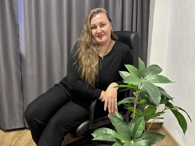 MonaLia with long, wavy brown hair, wearing black clothes, sitting in a chair, smiling, hand on green potted plant, wooden floor, gray curtains.