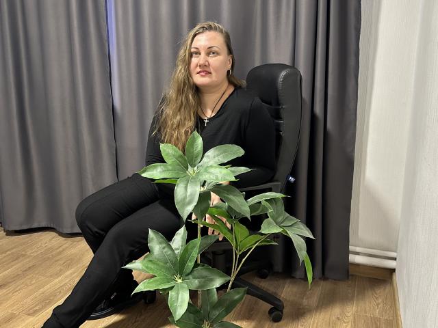 MonaLia with long blonde hair, black clothes, sitting on office chair, holding green plant with large leaves, wooden floor, grey curtains, black shoes, slightly smiling, relaxed.