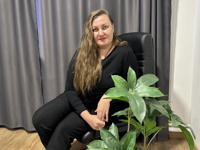 MonaLia with long brown hair, black clothes, sitting in chair, smiling, hands on knee, next to green potted plant, wooden floor, gray curtains. Her ass is slightly visible.