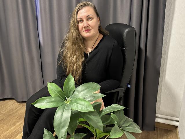MonaLia with long brown hair, black clothes, sitting in chair, holding green plant, wooden floor, gray curtains, black leather chair, calm expression, slight smile, medium tits, fair skin.
