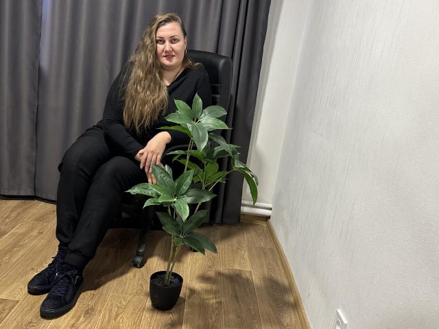 MonaLia with long brown hair, black clothes, black shoes, sitting on chair, smiling, next to green potted plant on wooden floor, grey curtain behind.
