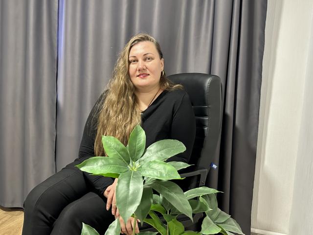 MonaLia with long blonde hair, black clothes, sitting on chair, holding green plant, smiling, grey curtains in background, wooden floor, black office chair, white wall on right.