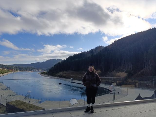 MonaLia stands on a balcony, wearing a brown jacket, black pants, and black shoes, with a cloudy sky and forested hills in the background. She has a small ass.