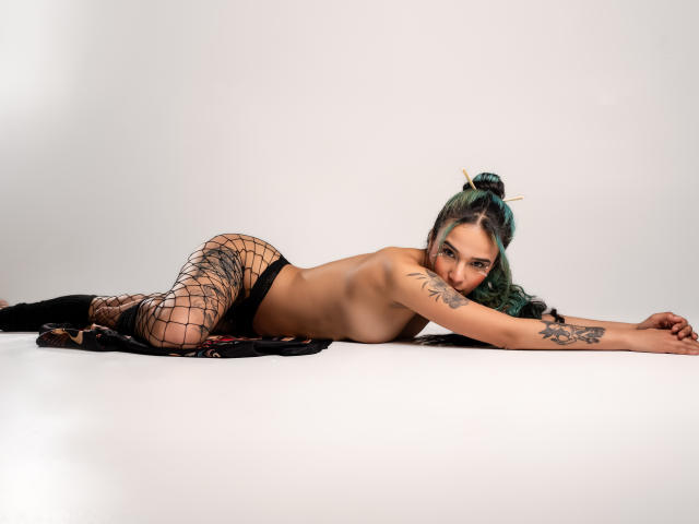 AliceHotBlue, topless, laying on her stomach, wearing black fishnet pants, with green hair in a bun, tattoo on her right arm, looking at the camera, smiling.