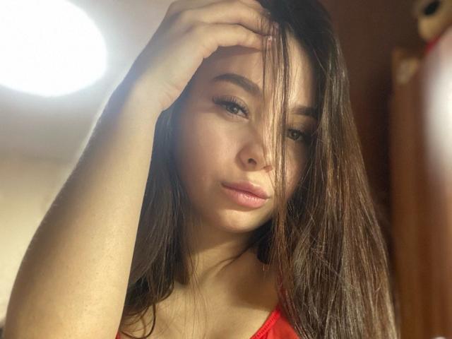 DramaVelvet with long brown hair, slightly parted lips, and a hand in her hair, wearing a red top, showing soft, subtle makeup, and a slight, sexy, confident expression.