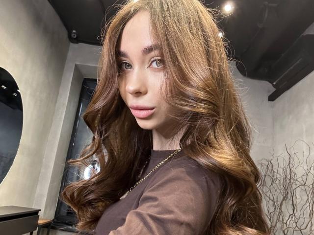 DramaVelvet has long, wavy brown hair, green eyes, and pink lips. She's wearing a brown shirt with a gold necklace, looking at the camera with a slight smile.