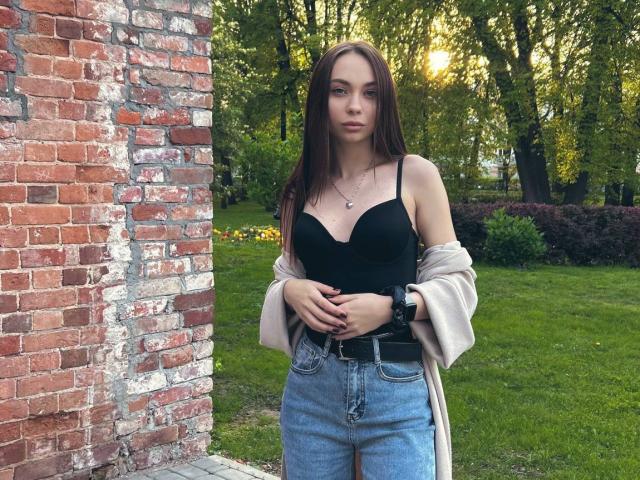 DramaVelvet with long brown hair, black strapless top, blue jeans, and beige coat, standing against a red brick wall, hands on waist, black watch, green grass behind.