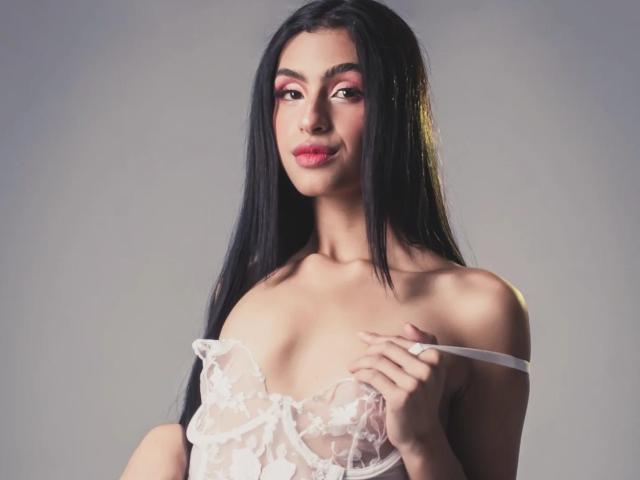 LilithDesire with long black hair, pink eyeshadow, and red lipstick, wearing a sheer white lace top, pulling one strap down, showing her small tits.