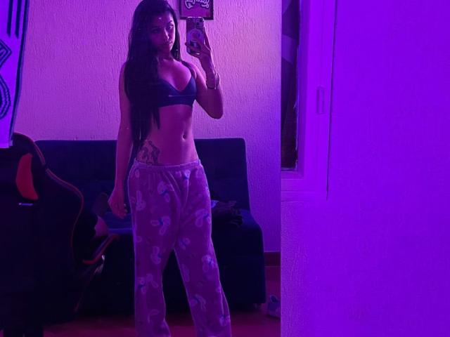 LilithDesire with long dark hair, black bra, and pink heart pajama pants, showing a tattoo on her right hip, taking a mirror selfie in purple-lit room.