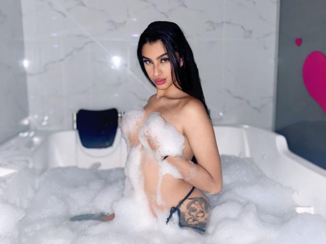 LilithDesire with long black hair, holding soapy bubbles over her small tits, wearing a black thong, tattoo on right hip, standing in a bubbly white bathtub.