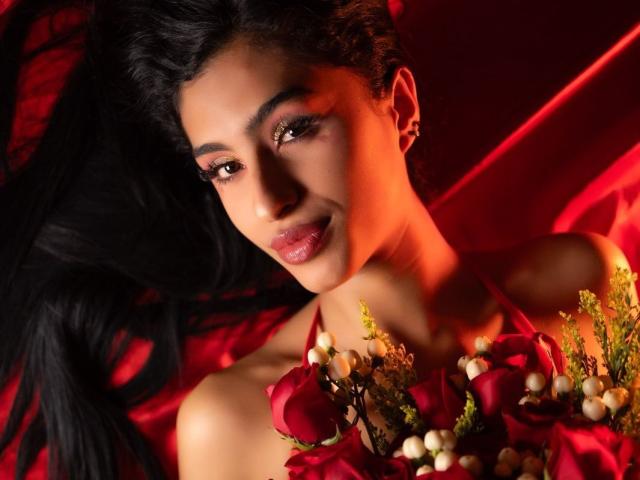 LilithDesire with dark hair, red eyes, and glossy lips, holding red roses and small white flowers, against a red satin background, looking seductively at the camera.