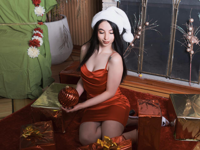 CamilleRios with black hair, wearing a Santa hat, red satin dress, holding an orange ornament, kneeling on red carpet, surrounded by gold-wrapped gifts. Boobs, ass, looking cute.