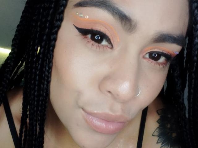 StellaBeckker with braided hair, orange eyeshadow, black eyeliner, small nose ring, pink lips, and a black tattoo on her right shoulder. Her eyes are looking slightly to the right.