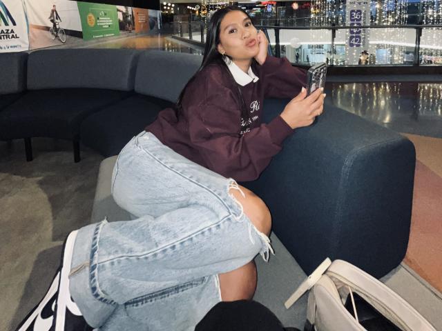 ThaliaAres, with dark hair and medium skin, leans on a blue chair, wearing a maroon hoodie and ripped blue jeans, holding a phone, with white sneakers and a bag.