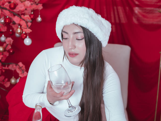 AnnFisher with long brown hair, white fluffy hat, white long-sleeve shirt, holding glass of water, bottle on table, red and pink Christmas tree ornaments, eyes closed, pink lips.