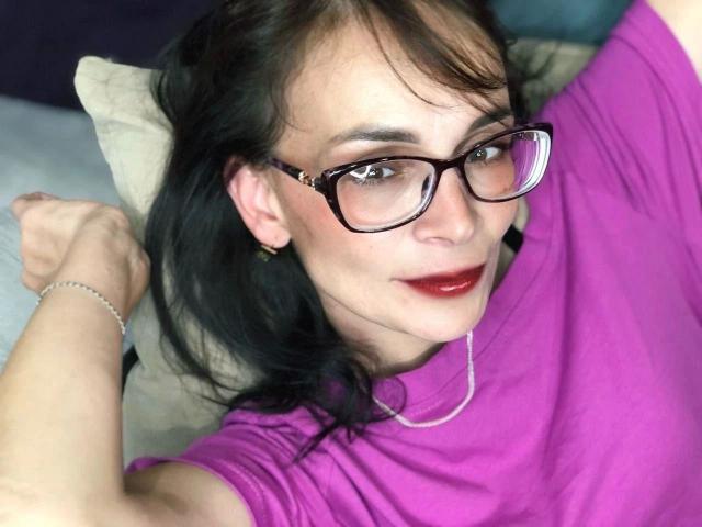 DarkOpiumX-hot with glasses, red lipstick, and purple shirt, lying on a pillow, raising arm, showing off small tits, black hair, and bracelet, looking at camera.