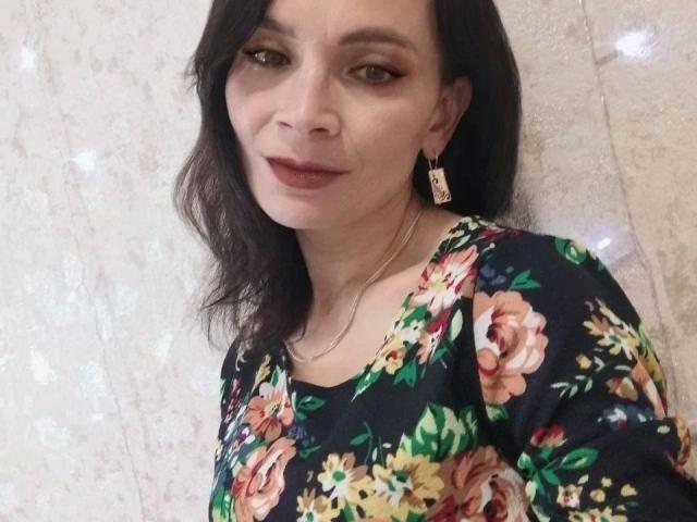 DarkOpiumX-hot with medium tits, wearing a floral dress, brown wavy hair, subtle makeup, and gold earrings, standing against a textured wall. She's smiling slightly.