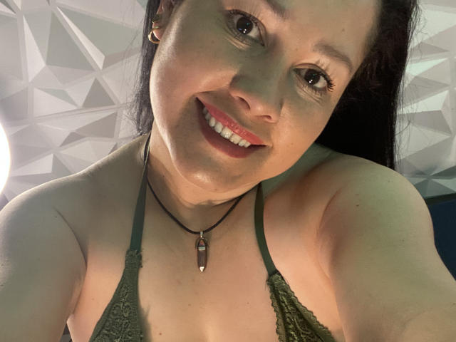MarianaSmitt with shiny skin, smiling, wearing a green lace halter top, black necklace with a gemstone, dark hair, and brown eyes, in a selfie. Boobs visible.