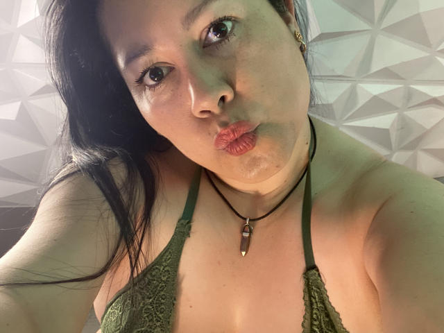 Close-up selfie of MarianaSmitt. She has light brown skin, dark straight hair, and is puckering her lips in a pout. She's wearing a green lace halter top that reveals her cleavage and a black choker with a small pendant. Her brown eyes are looking directly at the camera. The background has a geometric pattern in shades of gray. The image is well-lit, highlighting her facial features and the texture of the lace. The overall composition is centered and slightly tilted. The texture of her skin is smooth, and her expression is playful.