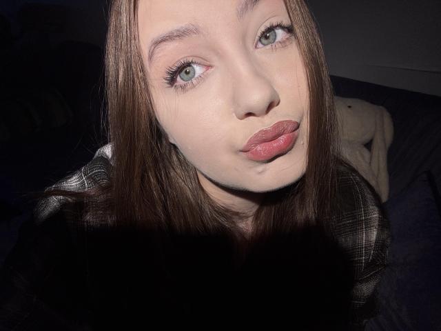 VannieAdelle has light green eyes, pink pouty lips, and straight brown hair. She's wearing a plaid shirt, with a shadowed black area in the foreground. Her ass is visible.