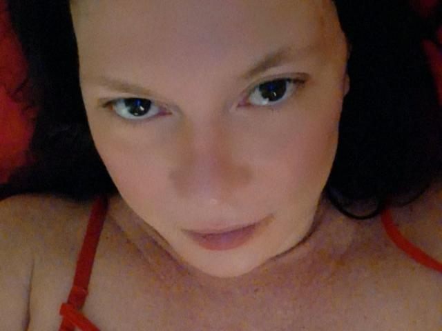 MeahBrunette with pale skin, dark eyes, and black hair, wearing a red strap, looking up with a subtle, sexy smile. Boobs visible, red strap over chest.