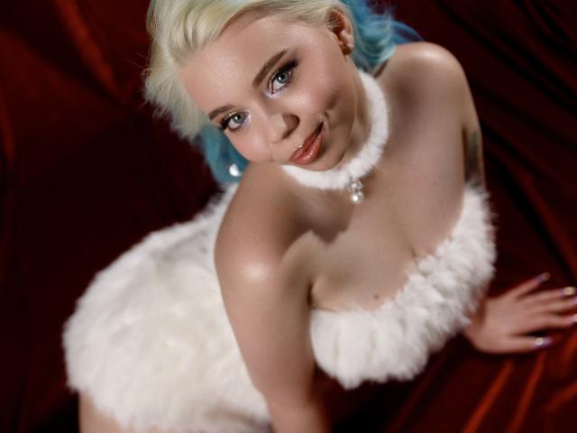 EnergyBurnny with blue hair, white fur dress, and pearl necklace, looking up with a smile. Her tits are small, and her ass is not visible. She's on red fabric.