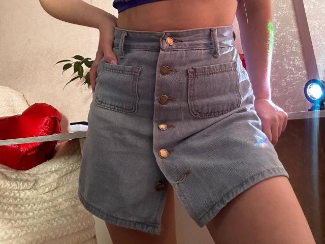 BalletSwan's denim skirt, buttoned down with copper buttons, showing her hands in pockets, wearing a blue crop top, standing with her ass slightly visible, in a casual setting.