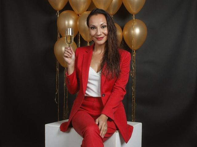LylyFrench, a medium-skinned woman with long brown hair, sits on a white cube. She's wearing a bright red suit with a white top, holding a champagne glass in her right hand. Gold balloons are behind her, adding a celebratory vibe. She has a confident, friendly smile. The image is well-lit, with a black background that makes her red suit and the gold balloons stand out. Her red lipstick matches her suit, and she looks relaxed and happy.