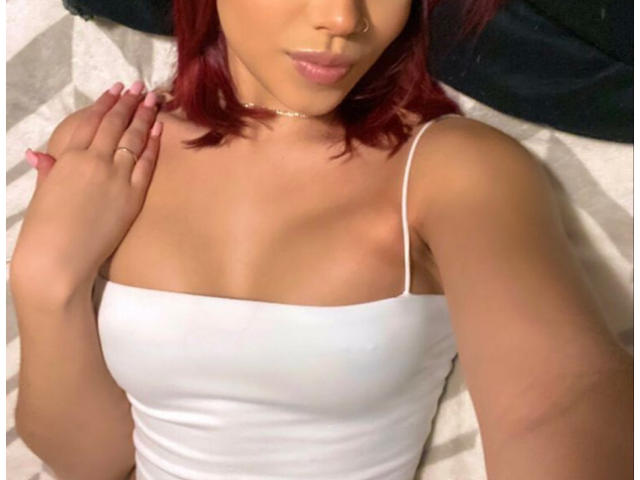 Roxelana with red hair, tan skin, and a nose ring, wearing a white tank top, showing her small tits and a choker necklace, with a hand on her shoulder.