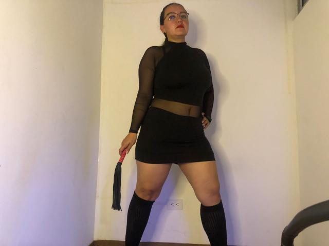 DominaRuby stands in a room, wearing a black sheer long-sleeve top, black mesh mini-skirt, black knee socks, holding a red and black flogger in her right hand.