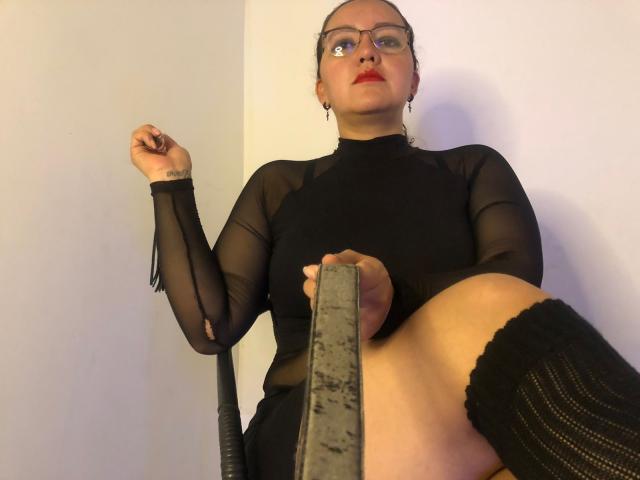 DominaRuby with glasses, red lipstick, and black sheer top, holding a grey chair, wearing black knee socks, looking up, tattoo on left wrist, white wall in background.