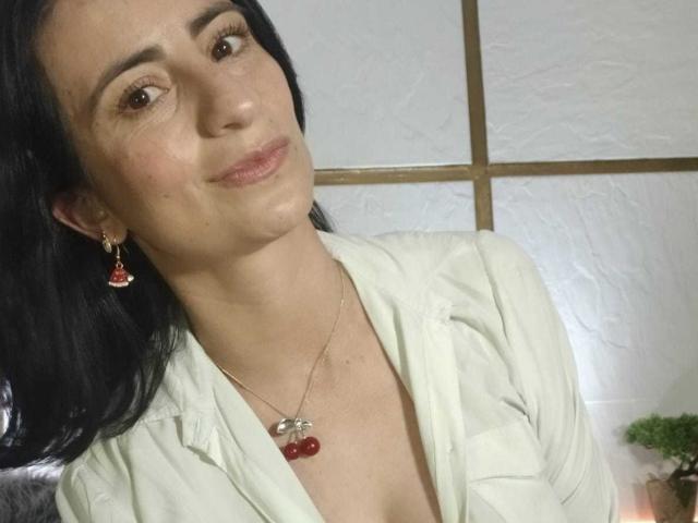 BrendaCantorel with dark hair, white shirt, red cherry necklace, and earrings, smiling slightly, showing her tits, pale skin, and brown eyes. She looks cute and confident.