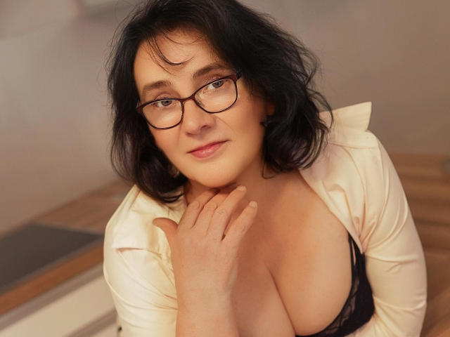 DorisMature has medium-length, wavy black hair and wears black-framed glasses. She has fair skin and is looking slightly upward with a subtle smile. She's dressed in a loose, white shirt with a black lace bra visible. Her right hand rests gently on her collarbone. The image has a warm, natural light, and her relaxed expression is prominent. The photo focuses on her upper body and face.