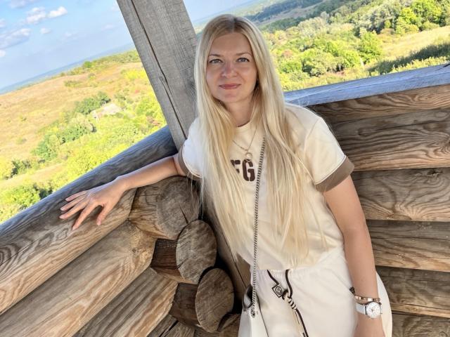 MelissaTyler with long blonde hair, white tee, and white shorts, leaning on wooden log structure, smiling, wearing a white watch, standing outside with green landscape in the background.