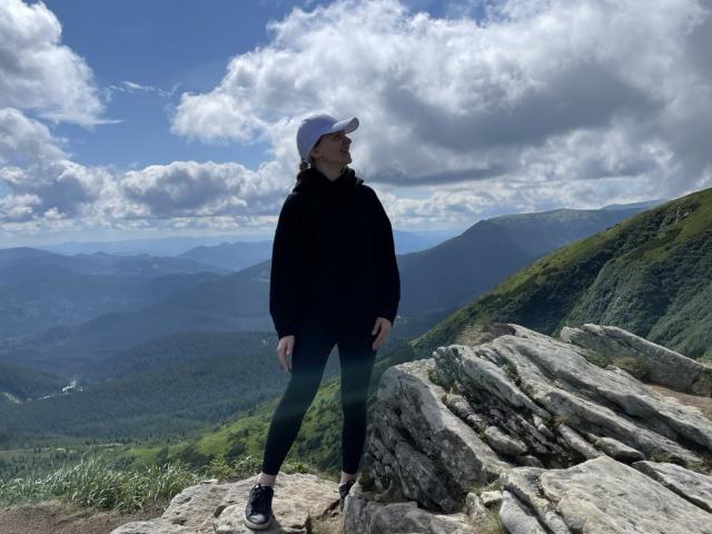 MelissaTyler stands on rocky mountain ledge, wearing black hoodie, black leggings, white cap, black shoes, looking to the right, with blue sky and clouds overhead.